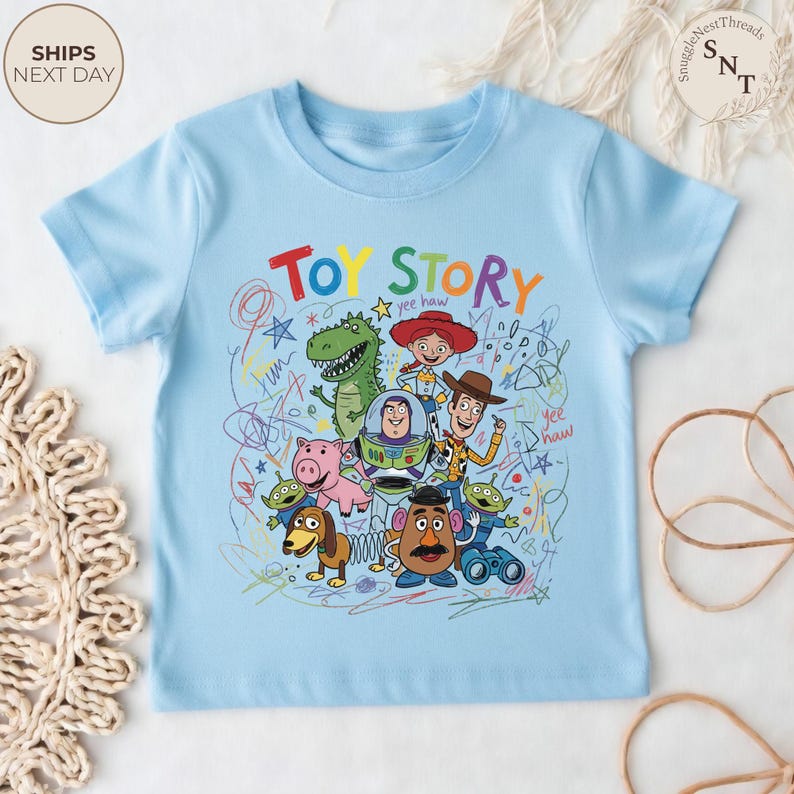 May include: Light blue t-shirt featuring the words "TOY STORY" in colorful letters. The shirt showcases a vibrant graphic of characters from the animated film, including Woody, Buzz Lightyear, and Rex, with scribbled background details.