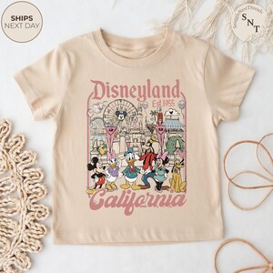 May include: A beige short-sleeve t-shirt featuring a graphic print of Disneyland California with the text Disneyland Est. 1955 California. The illustration includes Mickey Mouse, Minnie Mouse, Donald Duck, Daisy Duck, Goofy, and Pluto.