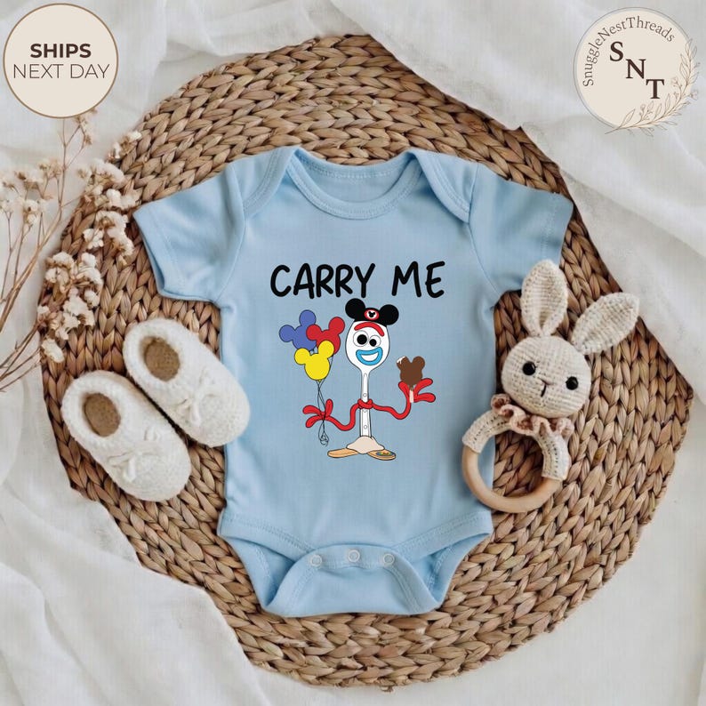 May include: A light blue baby onesie with the text "CARRY ME" and a cartoon character design. The design features a spoon with a face, holding balloons and a chocolate bar. The onesie is surrounded by white baby shoes and a bunny rattle.
