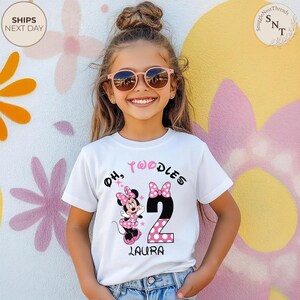 Puede incluir: Una camiseta blanca con un gr&aacute;fico de Minnie Mouse, el texto "Oh, Twoodles", el n&uacute;mero 2 con un dise&ntilde;o de lunares rosas y el nombre "Laura". La camiseta la lleva una ni&ntilde;a peque&ntilde;a con gafas de sol.