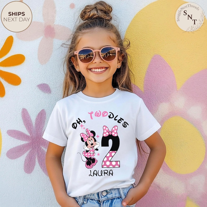 Puede incluir: Una camiseta blanca con un gr&aacute;fico de Minnie Mouse, el texto "Oh, Twoodles", el n&uacute;mero 2 con un dise&ntilde;o de lunares rosas y el nombre "Laura". La camiseta la lleva una ni&ntilde;a peque&ntilde;a con gafas de sol.