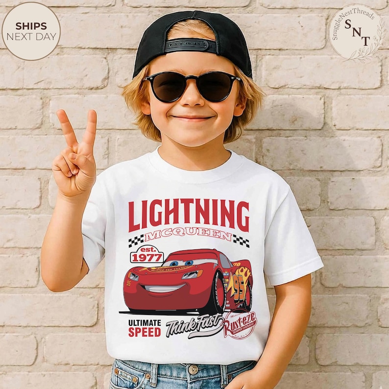 Op de afbeelding: Wit T-shirt met een Lightning McQueen-afbeelding uit de film Cars. Het ontwerp bevat de tekst "LIGHTNING MCQUEEN" boven de auto-afbeelding, met "est. 1977" en "ULTIMATE SPEED Think Fast Rusteze" eronder. Het T-shirt wordt door een kind gedragen.