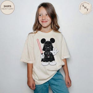 May include: A cream-colored t-shirt featuring a cartoon character mashup of Darth Vader and Mickey Mouse, holding a pink lightsaber. The character is in black and white with a red lightsaber. The shirt has the text "SnuggleNestThreads" and "SHIPS NEXT DAY".