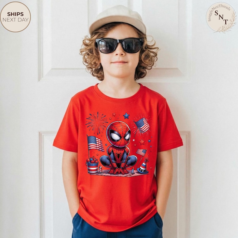 May include: A bright red short-sleeved t-shirt featuring a graphic of a cartoon spider-themed character surrounded by American flags, fireworks, and patriotic party decorations. The design includes red, white, and blue colors throughout the print.