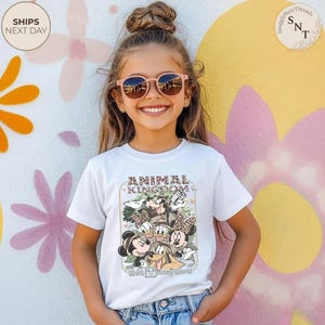 Pu&ograve; includere: T-shirt bianca con grafica colorata di personaggi Disney, testo "ANIMAL KINGDOM" e "Walt Disney World". La persona indossa occhiali da sole rosa. Sfondo colorato.
