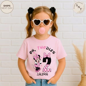 Puede incluir: Camiseta rosa para ni&ntilde;os peque&ntilde;os con un gr&aacute;fico de Minnie Mouse, el texto "Oh, Twoodles 2 Laura" y un n&uacute;mero 2 con lunares. El ni&ntilde;o lleva gafas de sol en forma de coraz&oacute;n y tiene lazos negros para el pelo.