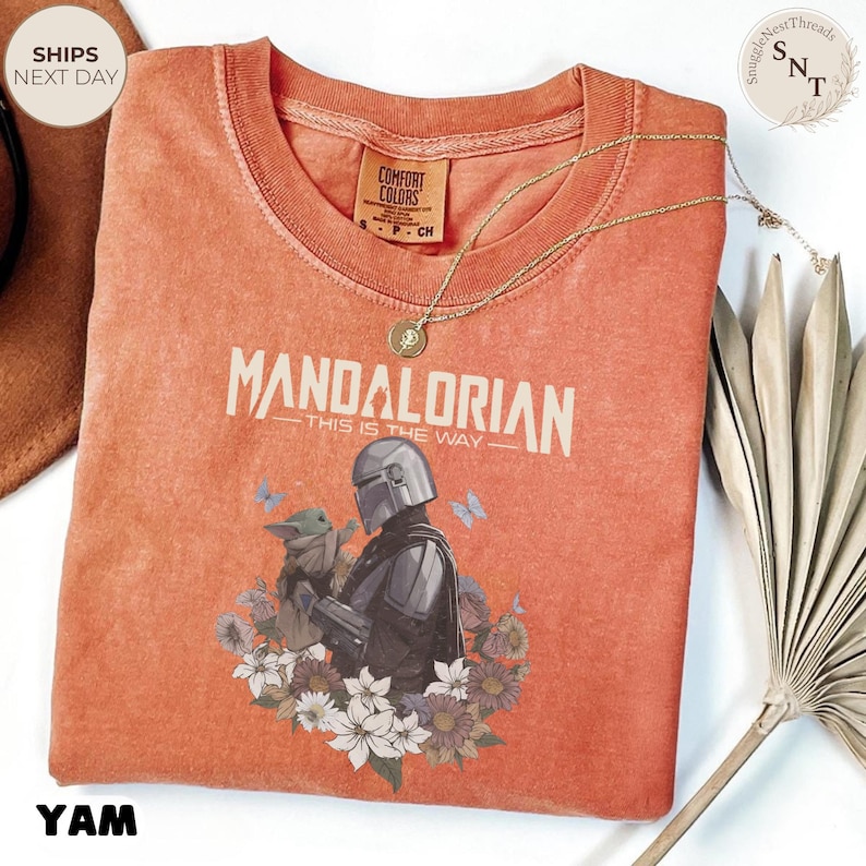 Puede incluir: Una camiseta de manga corta color naranja quemado con un gr&aacute;fico del Mandaloriano y Grogu rodeados de flores y mariposas. El texto dice Mandalorian This Is The Way. La camiseta tiene cuello redondo y un acabado lavado estilo vintage.
