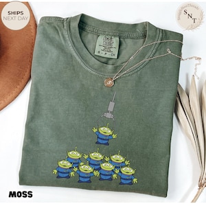 May include: Moss green t-shirt featuring a graphic of the Little Green Men from Toy Story being lifted by a claw. The shirt has a crew neck and short sleeves. The text "MOSS" is printed at the bottom.