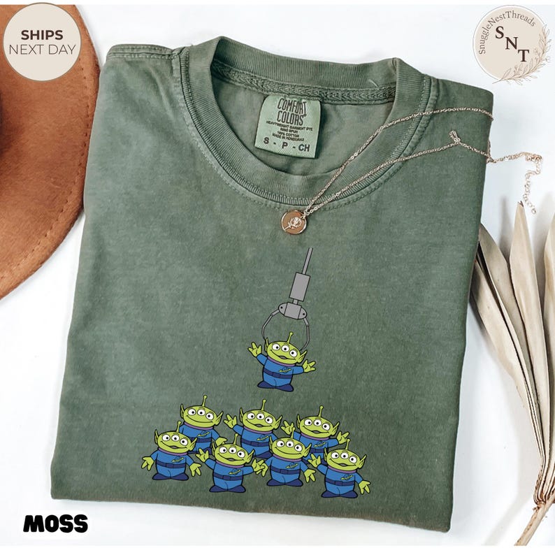 May include: Moss green t-shirt featuring a graphic of the Little Green Men from Toy Story being lifted by a claw. The shirt has a crew neck and short sleeves. The text "MOSS" is printed at the bottom.