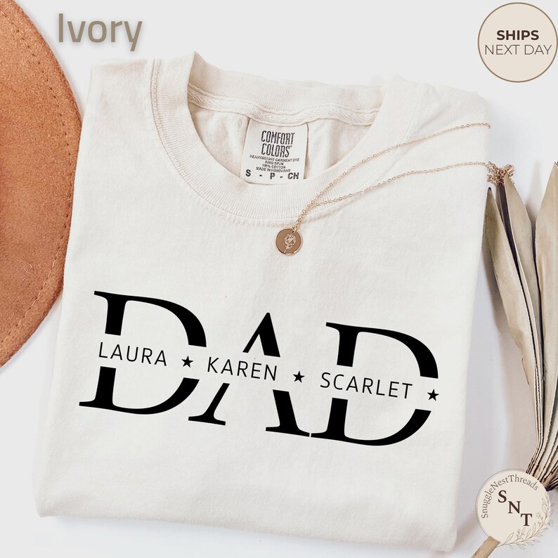 Puede incluir: Camiseta de color marfil con la palabra "DAD" en letras negras grandes. Debajo de la palabra "DAD" est&aacute;n los nombres "LAURA", "KAREN" y "SCARLET" separados por estrellas. La camiseta tiene cuello redondo y mangas cortas.