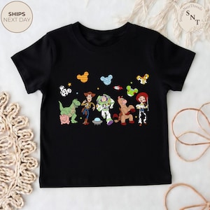 May include: Black t-shirt featuring a colorful graphic of characters from the Toy Story movie. The design includes Woody, Buzz Lightyear, Jessie, and other characters. The shirt has a crew neck and short sleeves.