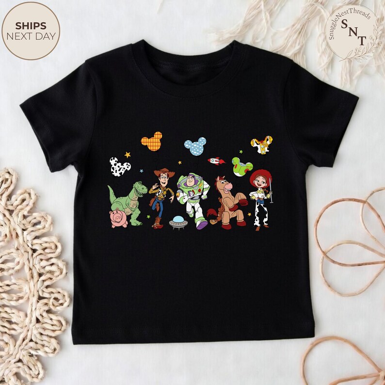 May include: Black t-shirt featuring a colorful graphic of characters from the Toy Story movie. The design includes Woody, Buzz Lightyear, Jessie, and other characters. The shirt has a crew neck and short sleeves.