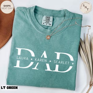 Puede incluir: Una camiseta Comfort Colors verde claro con la palabra "DAD" en letras blancas grandes, con los nombres LAURA, KAREN y SCARLET debajo. Un collar dorado con un colgante descansa sobre la camiseta. La camiseta est&aacute; etiquetada como "LT GREEN".