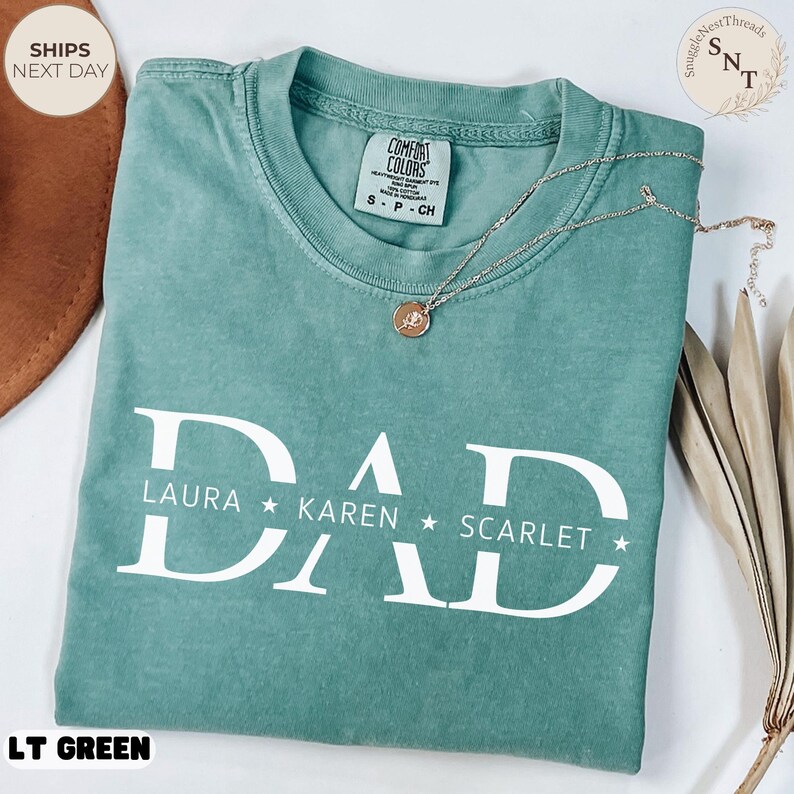 Puede incluir: Una camiseta Comfort Colors verde claro con la palabra "DAD" en letras blancas grandes, con los nombres LAURA, KAREN y SCARLET debajo. Un collar dorado con un colgante descansa sobre la camiseta. La camiseta est&aacute; etiquetada como "LT GREEN".