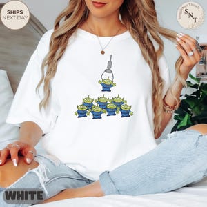 May include: White t-shirt featuring a graphic of green aliens from a claw machine. The aliens are in blue uniforms. The text "WHITE" is at the bottom left. The shirt has the text "SHIPS NEXT DAY" on the top left and the logo "SnuggleNestThreads" on the top right.