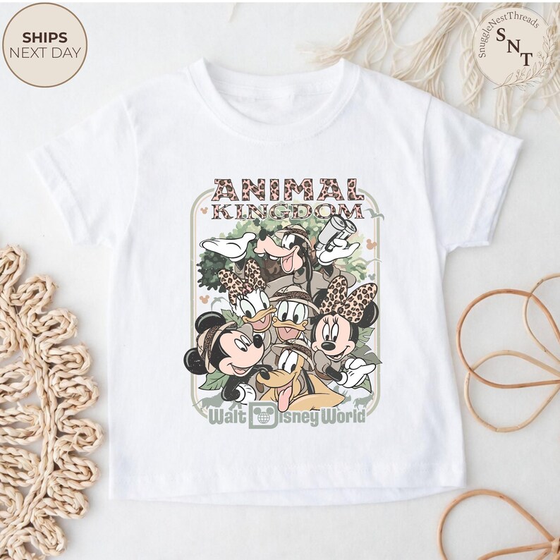 Pu&ograve; includere: T-shirt bianca con una grafica di Topolino, Minnie, Paperino, Paperina, Pippo e Pluto in tenuta da safari. Il testo "ANIMAL KINGDOM" e "Walt Disney World" sono anch'essi visualizzati sulla maglietta.