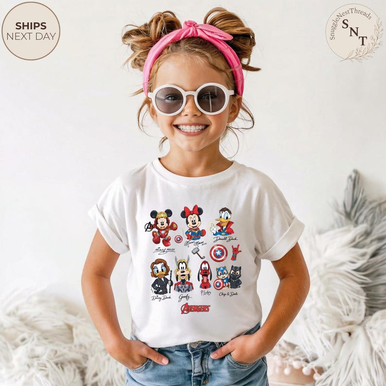 May include: A white t-shirt featuring Disney characters in superhero costumes. The shirt includes Mickey Mouse, Minnie Mouse, and Donald Duck, along with other characters and the word "Avengers". The child is wearing sunglasses and a pink headband.