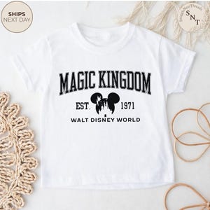 May include: White t-shirt with a black graphic that reads "MAGIC KINGDOM" above "EST. 1971" and "WALT DISNEY WORLD". The graphic includes a silhouette of Mickey Mouse ears and a castle.