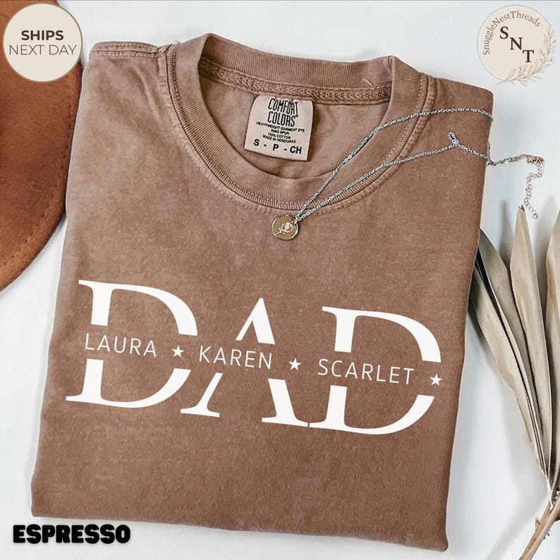 Puede incluir: Camiseta color espresso con la palabra "DAD" en letras blancas grandes, con los nombres Laura, Karen y Scarlet debajo. Un collar plateado con un colgante descansa sobre la camisa. La camiseta tiene una etiqueta Comfort Colors.