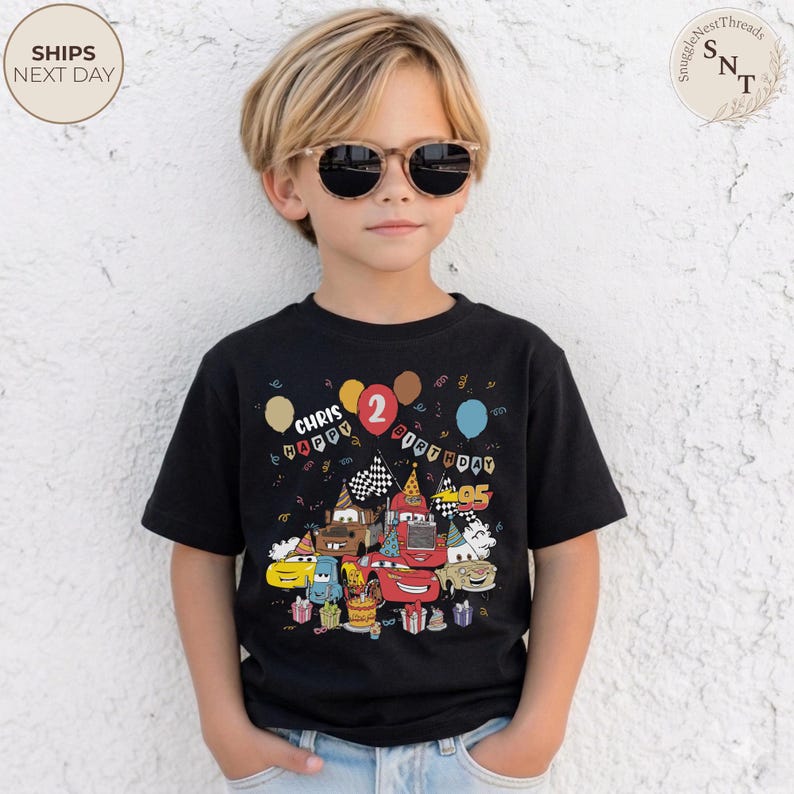 Puede incluir: Camiseta negra con un gr&aacute;fico colorido de coches de dibujos animados, globos y el texto "Chris Happy 2nd Birthday". El dise&ntilde;o incluye banderas de carreras y una tarta de cumplea&ntilde;os. El ni&ntilde;o lleva gafas de sol.