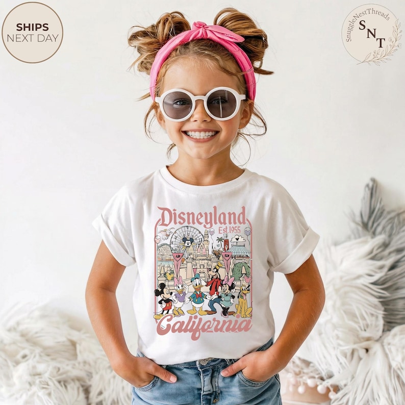 May include: A white short-sleeve t-shirt featuring a colorful graphic print of Disneyland California with Mickey Mouse, Minnie Mouse, Donald Duck, Daisy Duck, Goofy, and Pluto. The shirt is paired with a bright pink headband and round sunglasses.