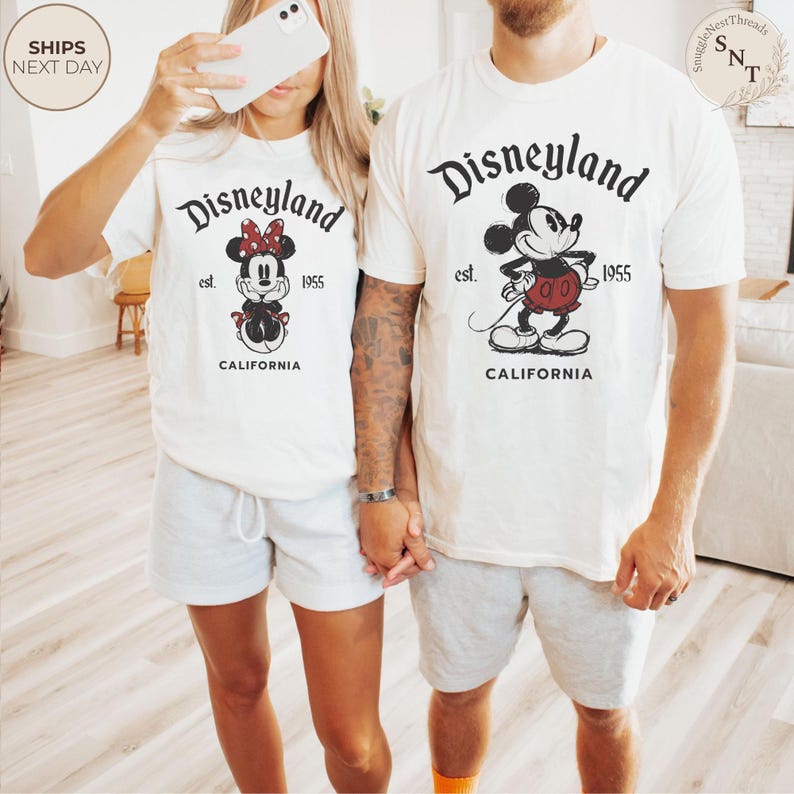 Pu&ograve; includere: Due magliette bianche sporco con il logo Disneyland, con Topolino e Minnie. Le magliette includono il testo "Disneyland est. 1955 California". I modelli indossano pantaloncini grigi. L'immagine &egrave; scattata in interni.