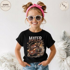 Puede incluir: Camiseta negra con un gr&aacute;fico de Mate de la pel&iacute;cula Cars, con el nombre "MATER" encima de la imagen. El dise&ntilde;o incluye detalles en marr&oacute;n, naranja y blanco. La ni&ntilde;a lleva una diadema rosa y gafas de sol blancas.