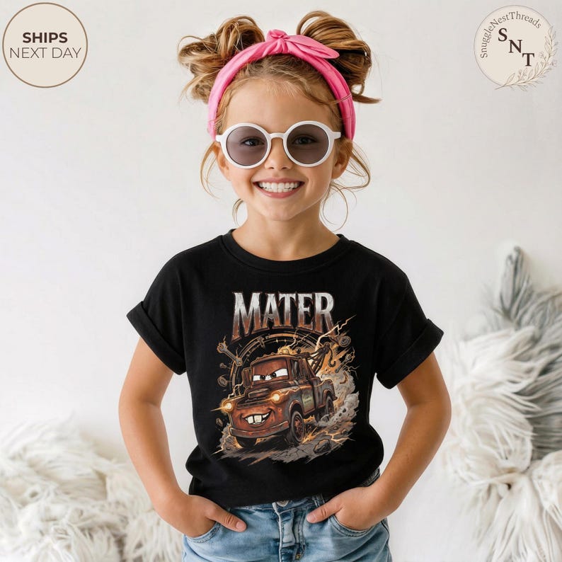 Puede incluir: Camiseta negra con un gr&aacute;fico de Mate de la pel&iacute;cula Cars, con el nombre "MATER" encima de la imagen. El dise&ntilde;o incluye detalles en marr&oacute;n, naranja y blanco. La ni&ntilde;a lleva una diadema rosa y gafas de sol blancas.