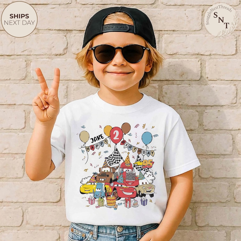 Puede incluir: Camiseta blanca con un gr&aacute;fico colorido de coches de dibujos animados, globos y el texto "JAKE HAPPY BIRTHDAY 2". El dise&ntilde;o incluye una tarta de cumplea&ntilde;os y regalos envueltos. El ni&ntilde;o lleva una gorra negra y gafas de sol.