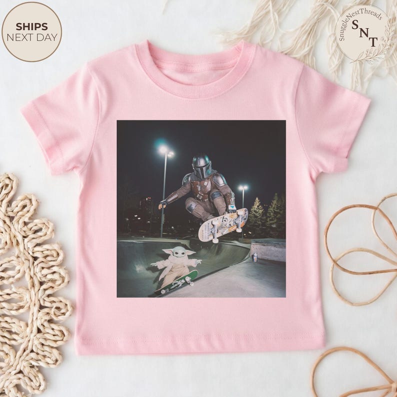 May include: A light pink t-shirt featuring a graphic of a character in a Mandalorian suit skateboarding. The image also includes a character from the Star Wars series, Grogu, skateboarding. The shirt has the text "SHIPS NEXT DAY" in a circle.