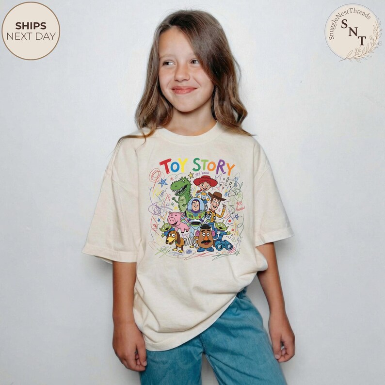 May include: A cream-colored t-shirt featuring the words "TOY STORY" in colorful letters. The shirt has a graphic of various Toy Story characters, including Woody, Buzz Lightyear, and Rex. The shirt is worn by a young person.
