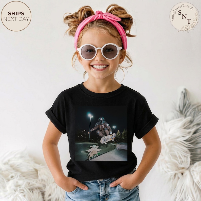 May include: Black t-shirt featuring a graphic of The Mandalorian and Grogu skateboarding at night. The image is centered on the shirt. The child is wearing sunglasses and a pink headband. The shirt has rolled-up sleeves.