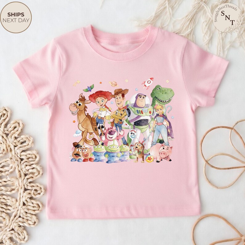 May include: A light pink t-shirt featuring a colorful graphic of characters from the Toy Story movie. The design includes Woody, Buzz Lightyear, Jessie, and other beloved characters. The shirt is made of a soft material and is suitable for children.