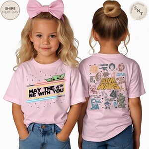May include: Pink Star Wars t-shirt with the text "May the 4th Be With You" and a graphic of Baby Yoda and lightsabers. The back of the shirt features Star Wars characters and quotes. The child is wearing blue jeans and a pink bow.