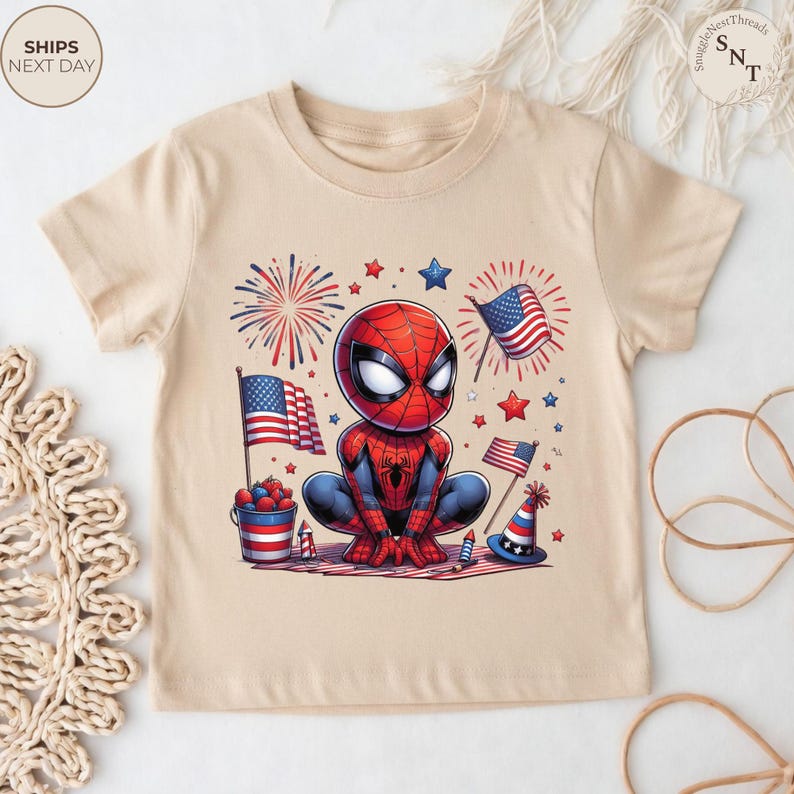 May include: A beige short-sleeve t-shirt featuring a graphic of a cartoon Spider-Man character surrounded by American flags, fireworks, stars, a bucket of berries, and a party hat. The design uses a red, white, and blue color palette.