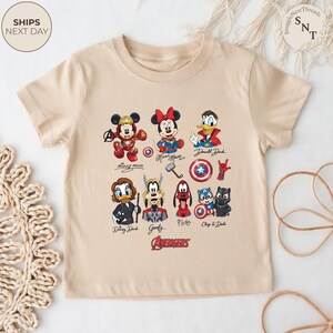 May include: A beige t-shirt featuring colorful illustrations of Disney characters as Avengers. The shirt includes Mickey Mouse, Minnie Mouse, Donald Duck, Daisy Duck, Goofy, Pluto, Chip & Dale, and the Avengers logo. The text "SHIPS NEXT DAY" is visible.