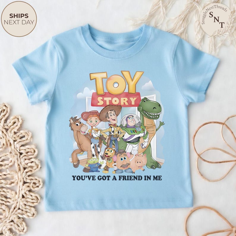 K&ouml;nnte beinhalten: Hellblaues T-Shirt mit dem Schriftzug "TOY STORY" in Gold und Rot, zusammen mit einer Grafik von Charakteren aus dem Film. Der Satz "YOU'VE GOT A FRIEND IN ME" ist darunter gedruckt. Kinderbekleidung.