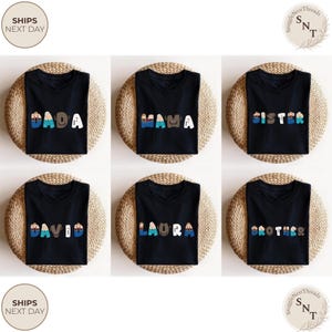 K&ouml;nnte beinhalten: Sechs schwarze T-Shirts mit verschiedenen Namen in bunten Buchstaben bedruckt: DADA, MAMA, SISTER, DAVID, LAURA und BROTHER. Jedes Shirt wird auf einer runden, gewebten Unterlage pr&auml;sentiert.