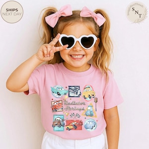 May include: A light pink t-shirt featuring a colorful graphic of various cartoon cars in a stamp-like design. The text "Radiator Springs" is printed in the center. The image also shows a young person wearing heart-shaped sunglasses and pink bows in their hair.