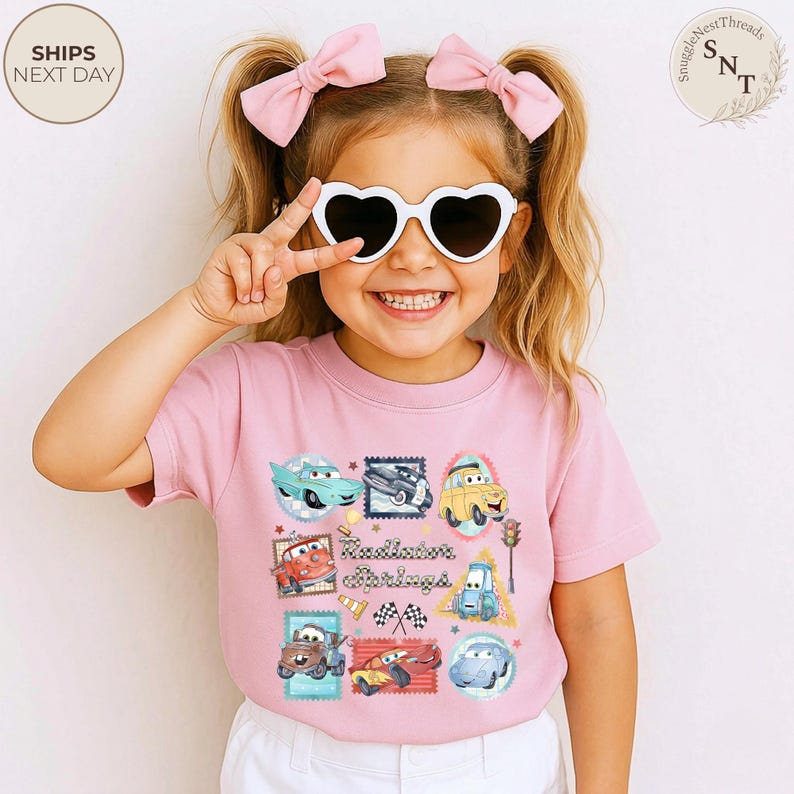 May include: A light pink t-shirt featuring a colorful graphic of various cartoon cars in a stamp-like design. The text "Radiator Springs" is printed in the center. The image also shows a young person wearing heart-shaped sunglasses and pink bows in their hair.