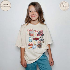 May include: A cream-colored t-shirt featuring a colorful Cars movie graphic. The design includes various cartoon cars, the word "Cars", and other decorative elements. The shirt is worn by a person.