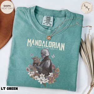 Puede incluir: Una camiseta verde claro con un gr&aacute;fico del Mandaloriano y Grogu rodeados de flores y mariposas. El texto dice Mandalorian This Is The Way. La camiseta tiene una etiqueta de Comfort Colors en el cuello y un collar de cadena dorada.