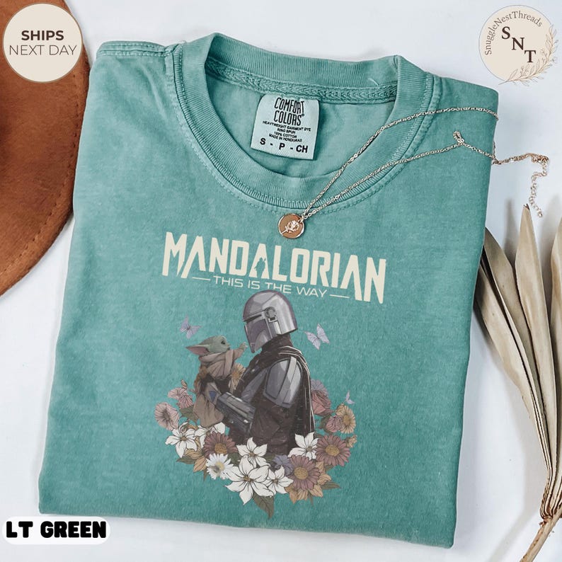 Puede incluir: Una camiseta verde claro con un gr&aacute;fico del Mandaloriano y Grogu rodeados de flores y mariposas. El texto dice Mandalorian This Is The Way. La camiseta tiene una etiqueta de Comfort Colors en el cuello y un collar de cadena dorada.