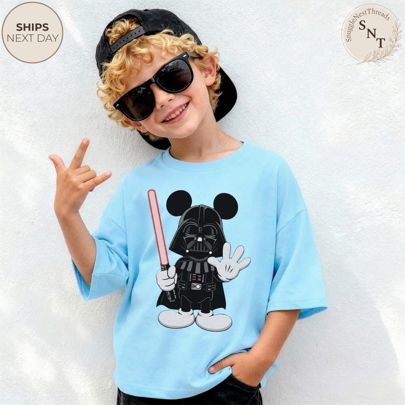 May include: Light blue t-shirt featuring a cartoon mashup of Darth Vader and Mickey Mouse. The design shows Darth Vader's helmet and cape, with Mickey Mouse's ears and gloves. The shirt also has a pink lightsaber.