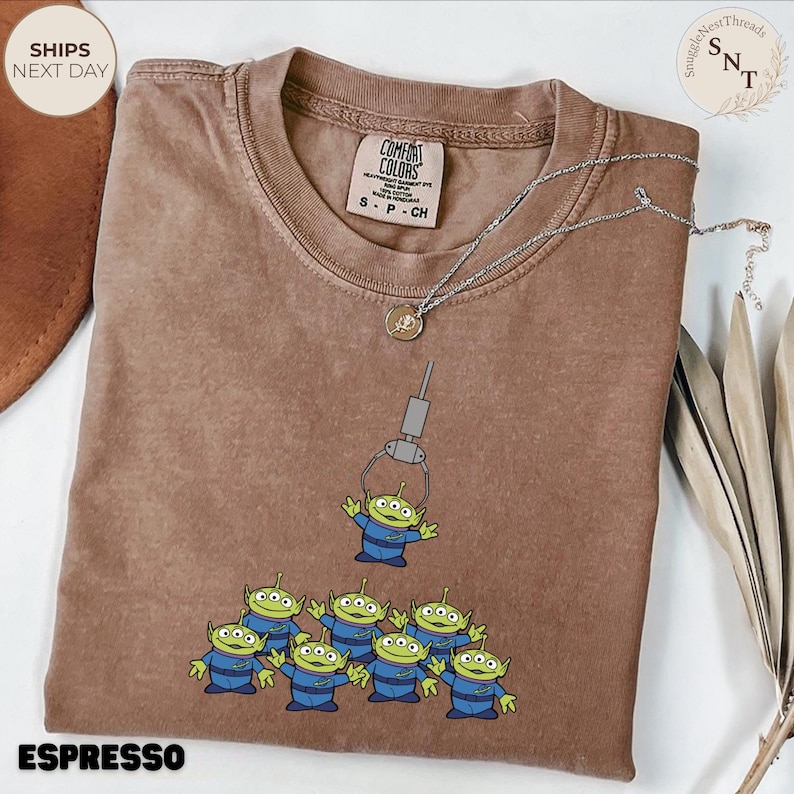 May include: Espresso-colored t-shirt featuring a cartoon claw machine lifting a green alien from a group of aliens. The shirt has a Comfort Colors tag and the word "ESPRESSO" at the bottom. A silver necklace with a pendant is draped on the shirt.