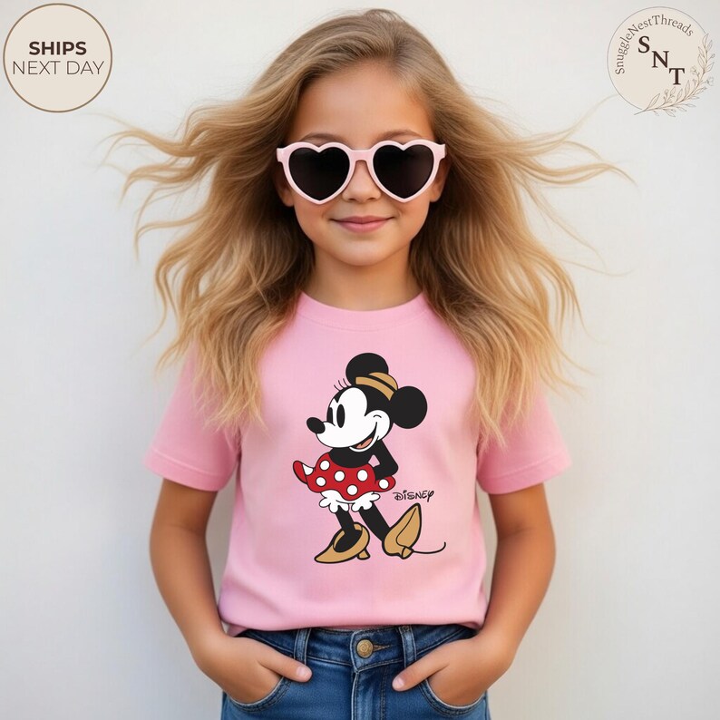 May include: A pink t-shirt featuring a Minnie Mouse graphic, paired with blue jeans. The Minnie Mouse design includes a black and white face, a red and white polka dot skirt, and gold shoes. The image also shows a young person wearing heart-shaped sunglasses.