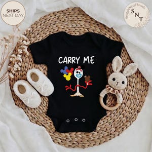 May include: Black baby onesie with the text "CARRY ME" and a cartoon character design. The design includes a spoon-fork character holding balloons and ice cream. The onesie is surrounded by white baby shoes and a stuffed bunny toy.