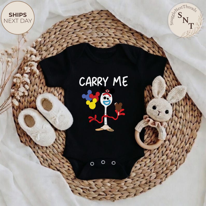 May include: Black baby onesie with the text "CARRY ME" and a cartoon character design. The design includes a spoon-fork character holding balloons and ice cream. The onesie is surrounded by white baby shoes and a stuffed bunny toy.