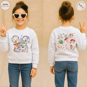 Puede incluir: Una sudadera blanca de manga larga con un gr&aacute;fico frontal de varios personajes animados dispuestos en forma de orejas de Mickey Mouse con un castillo y fuegos artificiales, y un gr&aacute;fico trasero con caras y nombres de personajes.