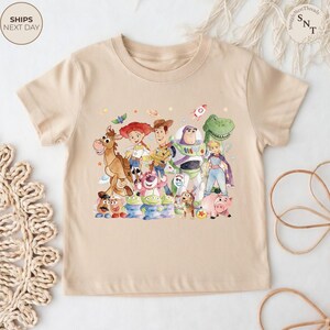 May include: A beige t-shirt featuring a colorful watercolor illustration of characters from the Toy Story movie franchise. The design includes Woody, Buzz Lightyear, Jessie, and other characters. The shirt has short sleeves and a crew neck.